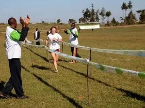 A female runner crossing the finish line in an outdoor race, with three officials or spectators nearby on a grassy field.