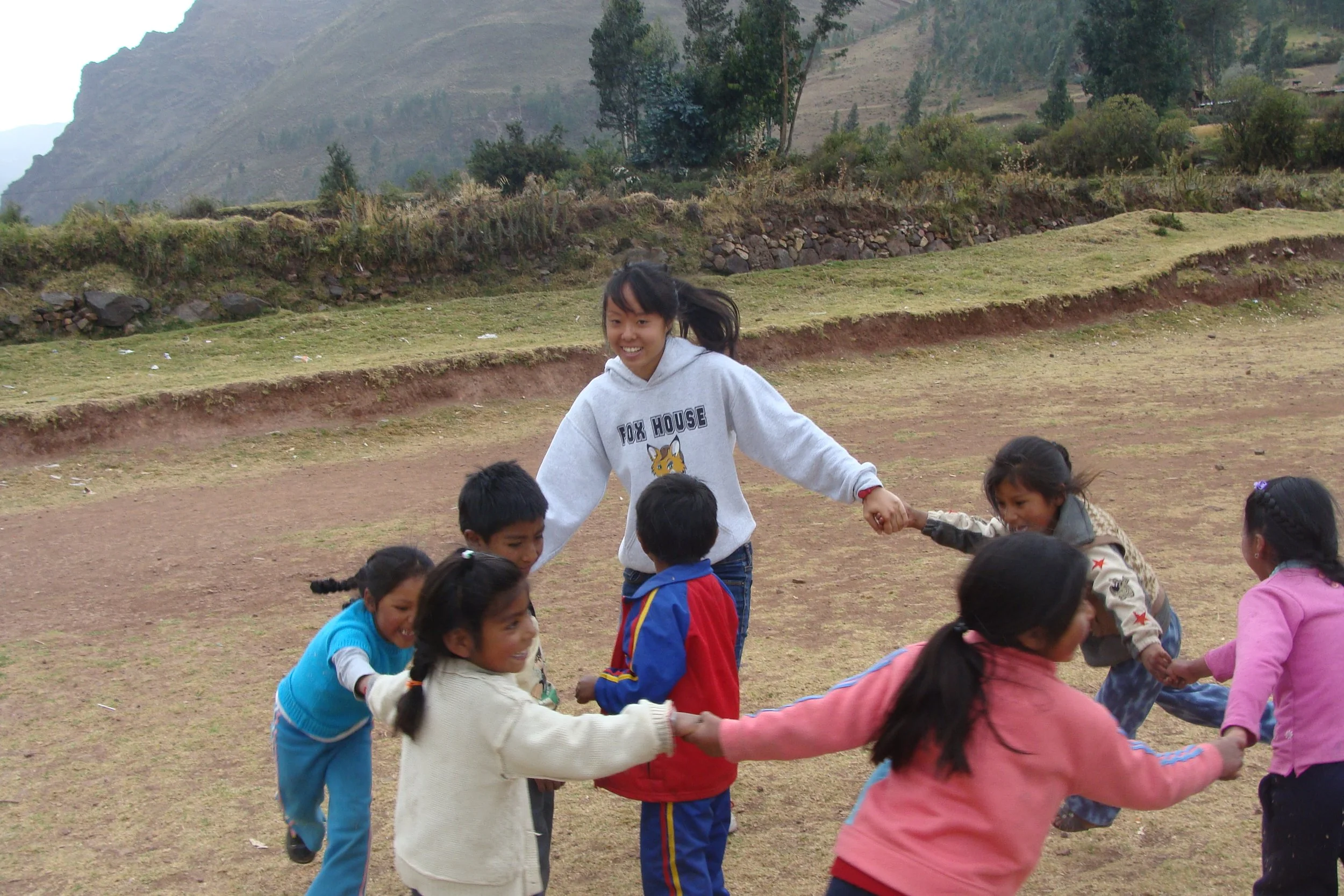 A young girl in a gray hoodie playing with children outdoors, holding hands in a circle on a grassy field with mountains and trees in the background.