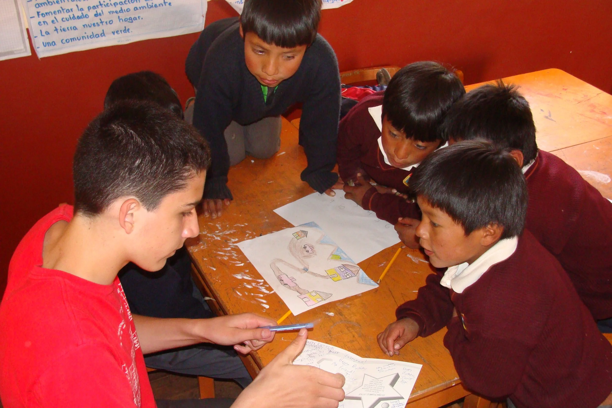 A group of young boys gathered around a table looking at drawings and a piece of paper, with some posters on the wall in the background.