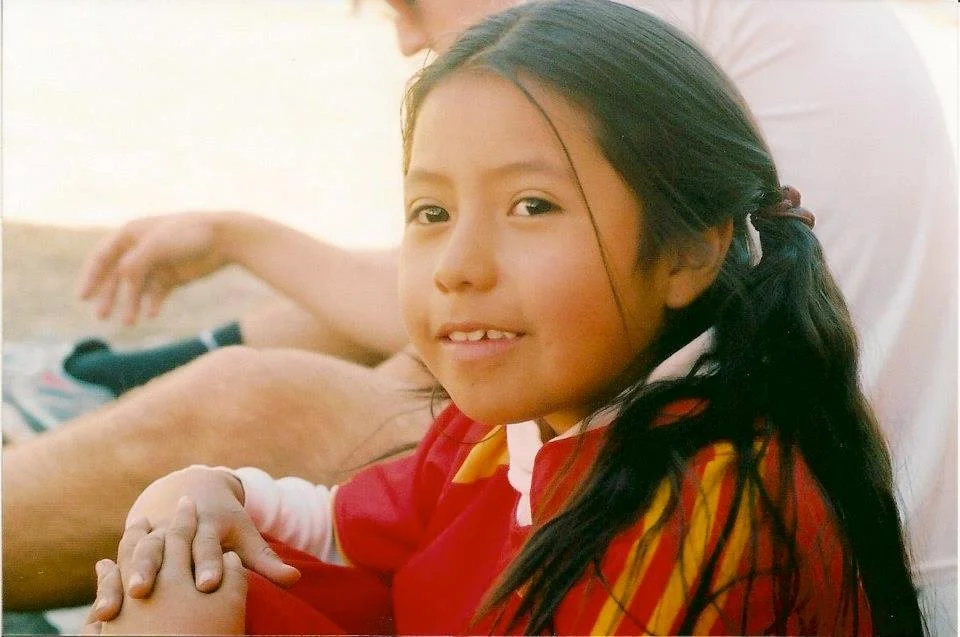 A young girl with long black hair and a red and yellow plaid shirt, smiling and holding a person's hand.