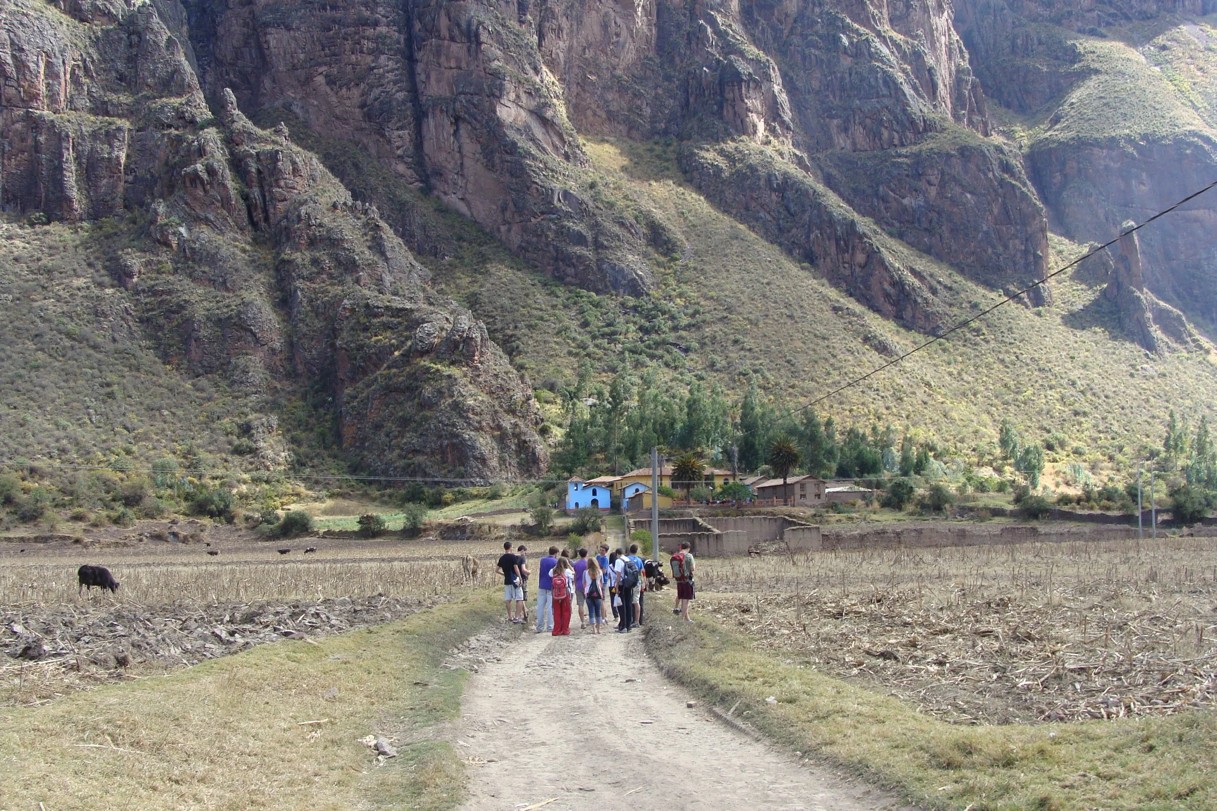 A group of people walking on a dirt path through a rural landscape with mountains in the background and a few houses near the base of the mountains.