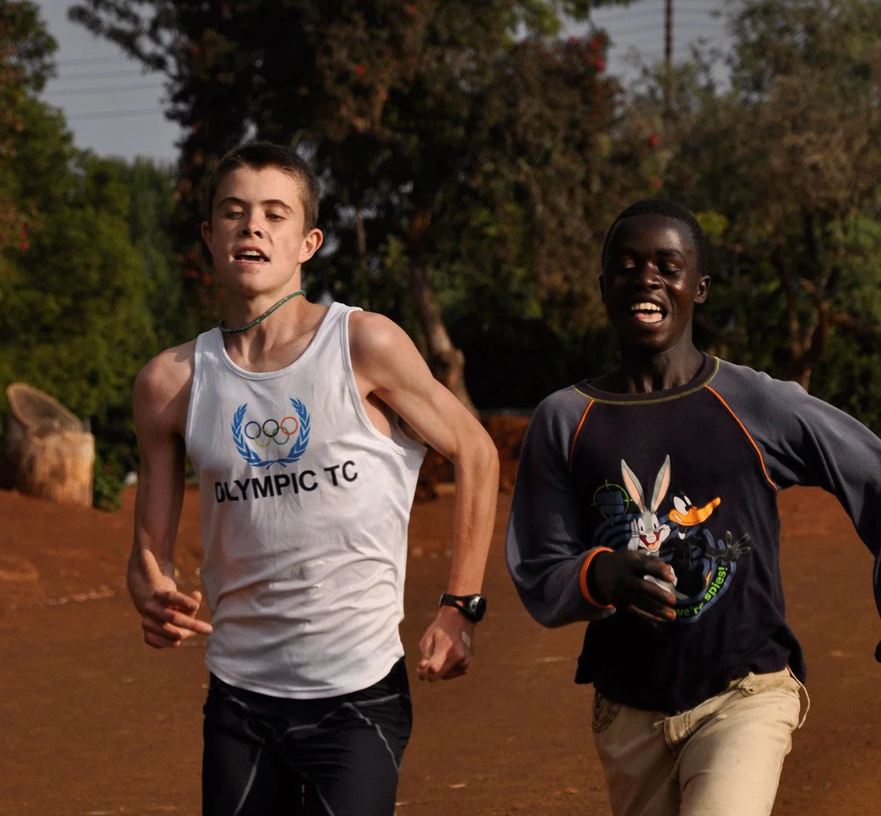 Two young male runners competing outdoors during a race, one wearing a white tank top with the Olympic logo, the other in a dark long-sleeve shirt with cartoon characters, both appearing focused and determined.