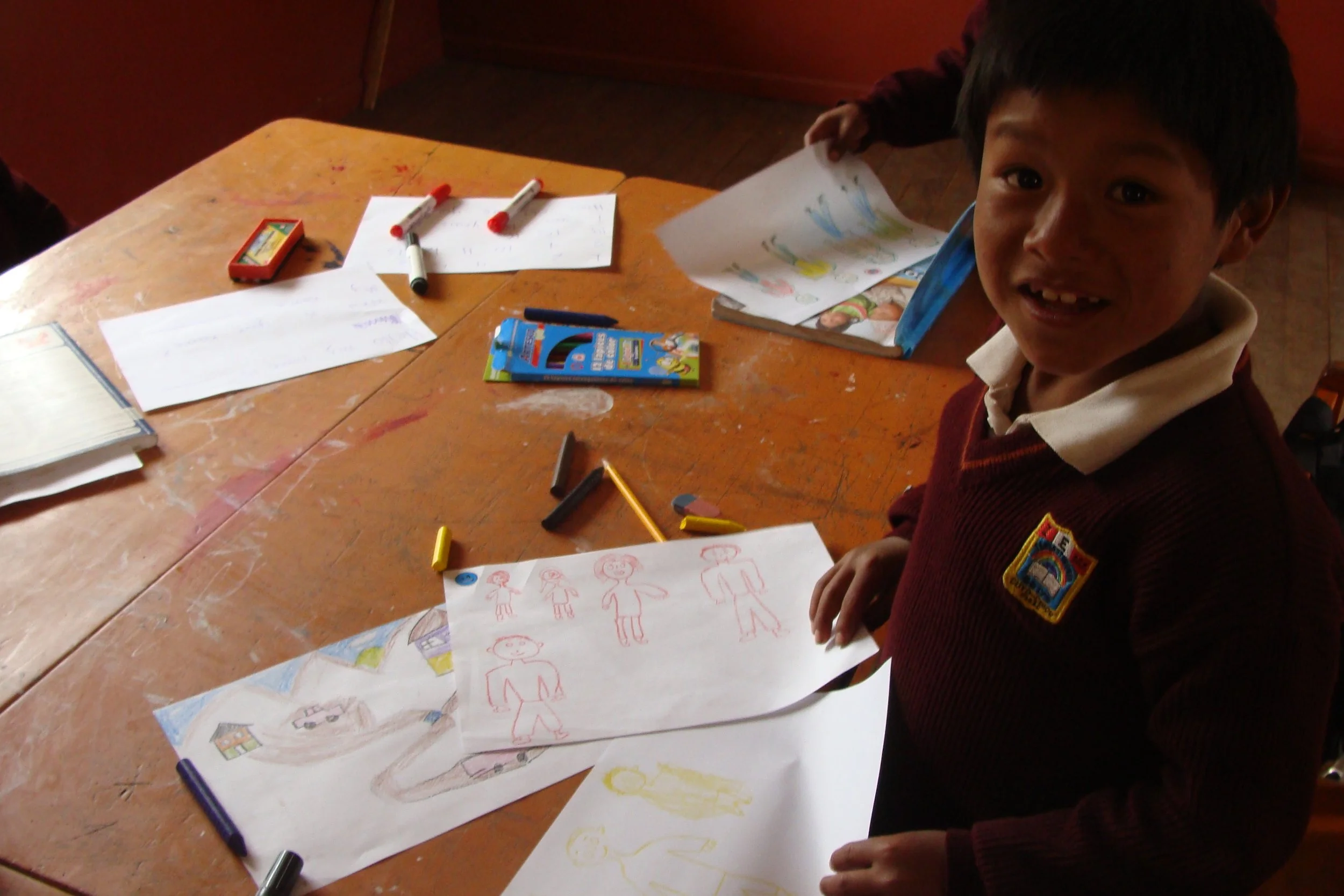 A young boy working on drawing pictures at a school desk with various papers, crayons, and markers scattered around.