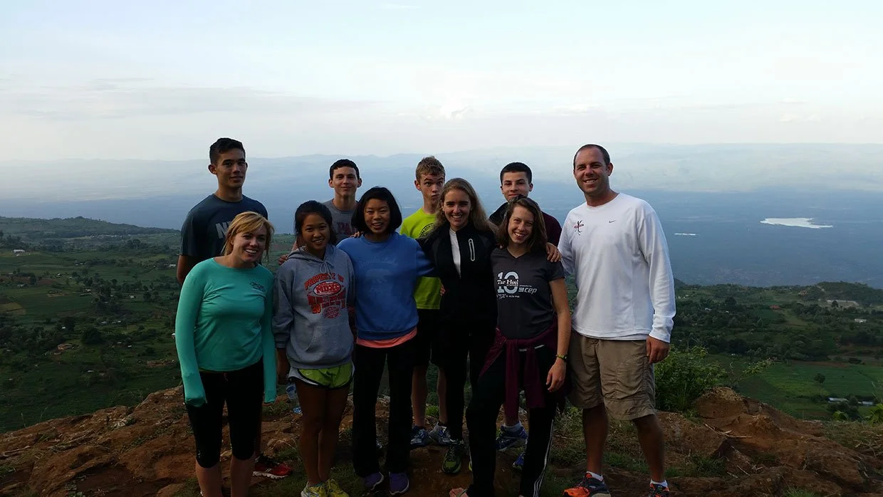 Group of ten people, including teenagers and adults, standing on a mountain with a landscape of green fields and a lake in the background.