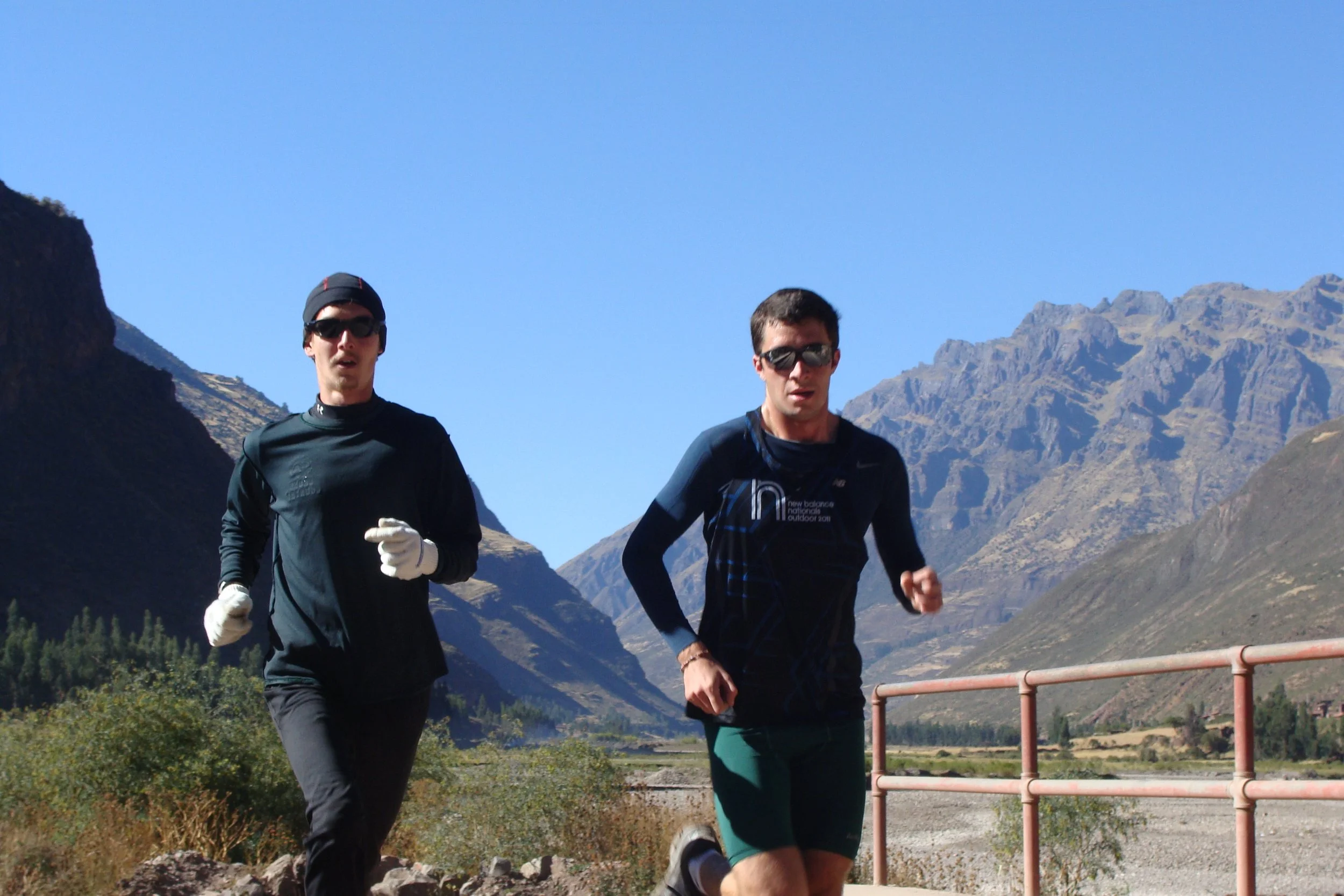 Two men running outdoors on a trail with mountains and blue sky in the background.