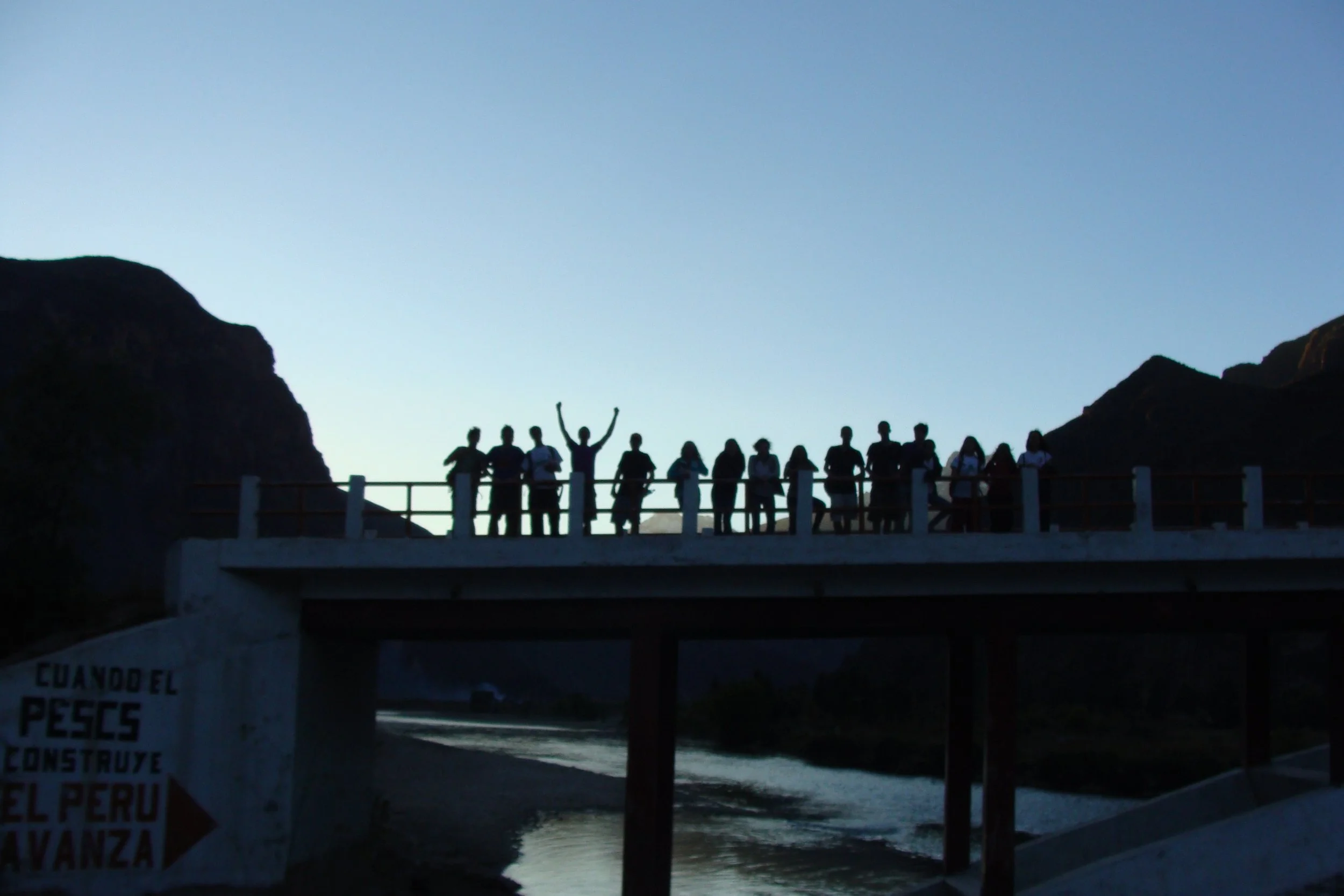 Group of people standing on a bridge silhouetted against a clear sky, with mountains in the background and a body of water below.