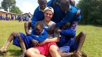 Young man sitting on grass surrounded by four boys in blue uniforms, with a schoolyard in the background.