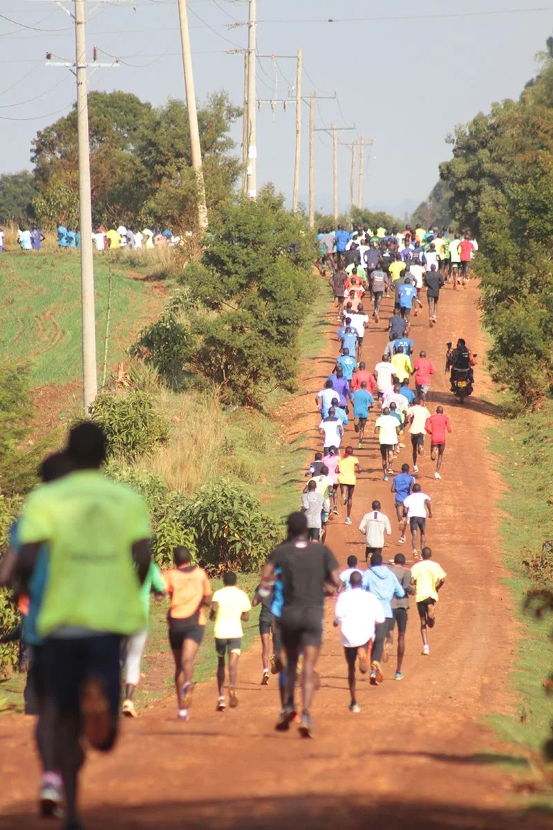 A large group of people, mainly running, participating in a long-distance race or marathon on a dirt road surrounded by greenery and power lines.