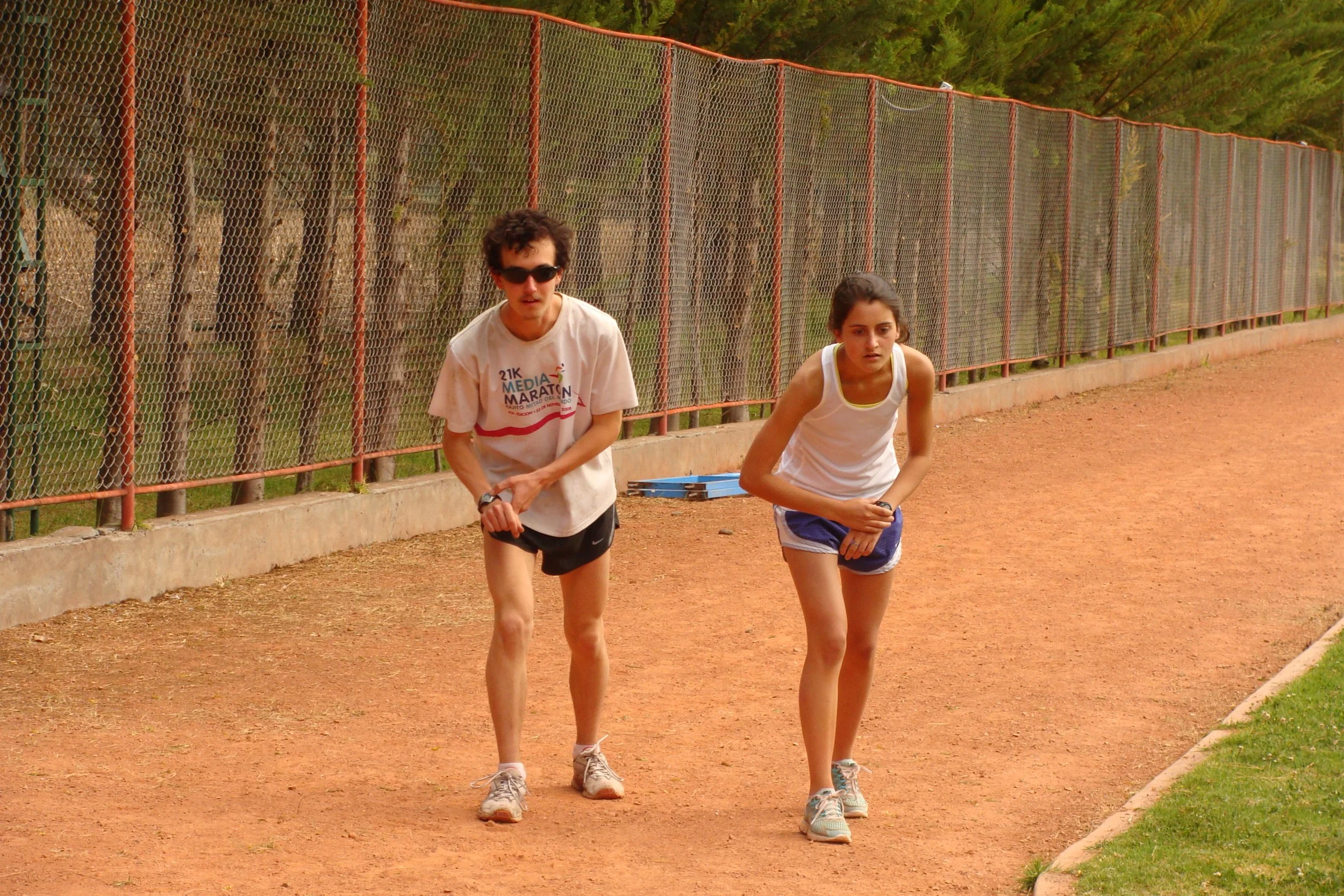 A man and a woman walking on a dirt track, with a chain-link fence and trees in the background.