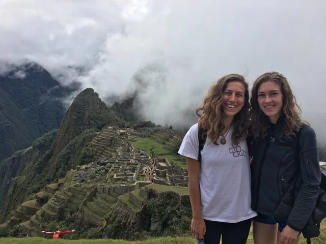 Two smiling women standing in front of Machu Picchu ruins with mountains and clouds in the background.