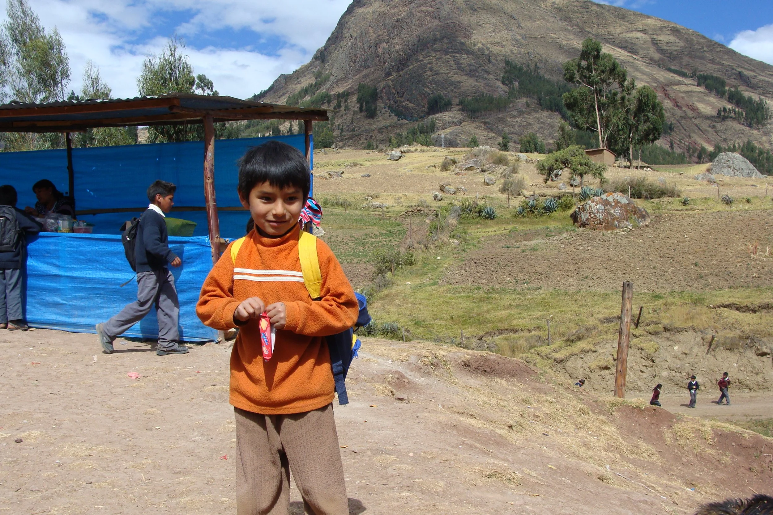 A young boy with a yellow backpack holding a candy, standing outdoors in a rural mountainous area with a small market stall behind him and other children walking in the background.