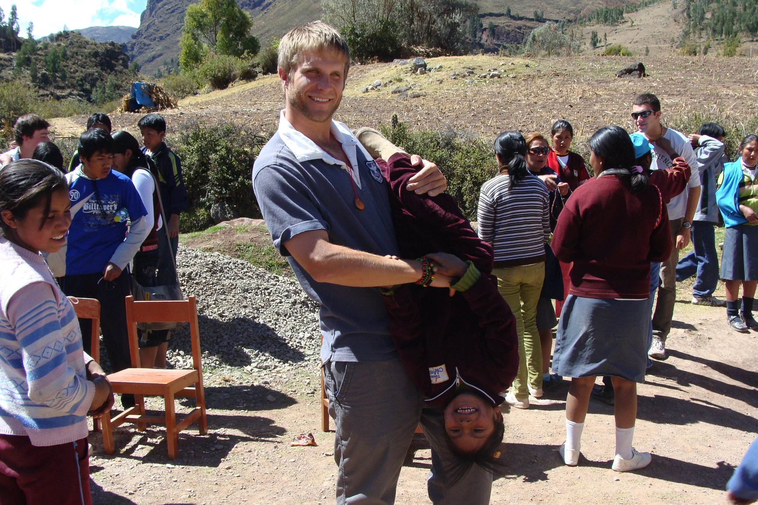 A man smiling while holding a young girl upside down in a crowded outdoor setting with mountains and trees in the background. Several children and adults are gathered, some looking at the camera and others engaged in conversation.