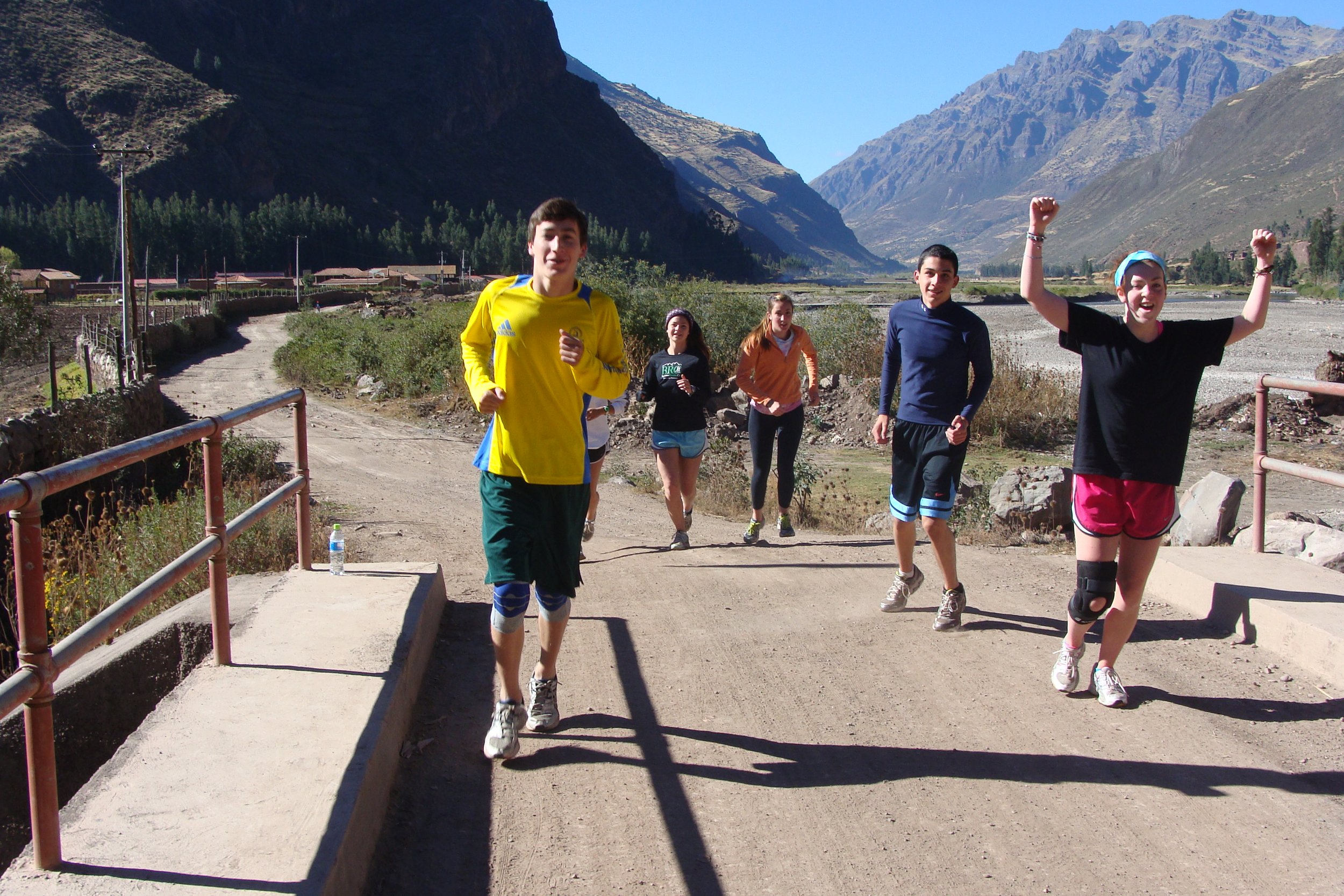 Group of six young people running outdoors on a dirt path in a mountainous area with blue sky, mountains, and trees in the background, some smiling and cheering.