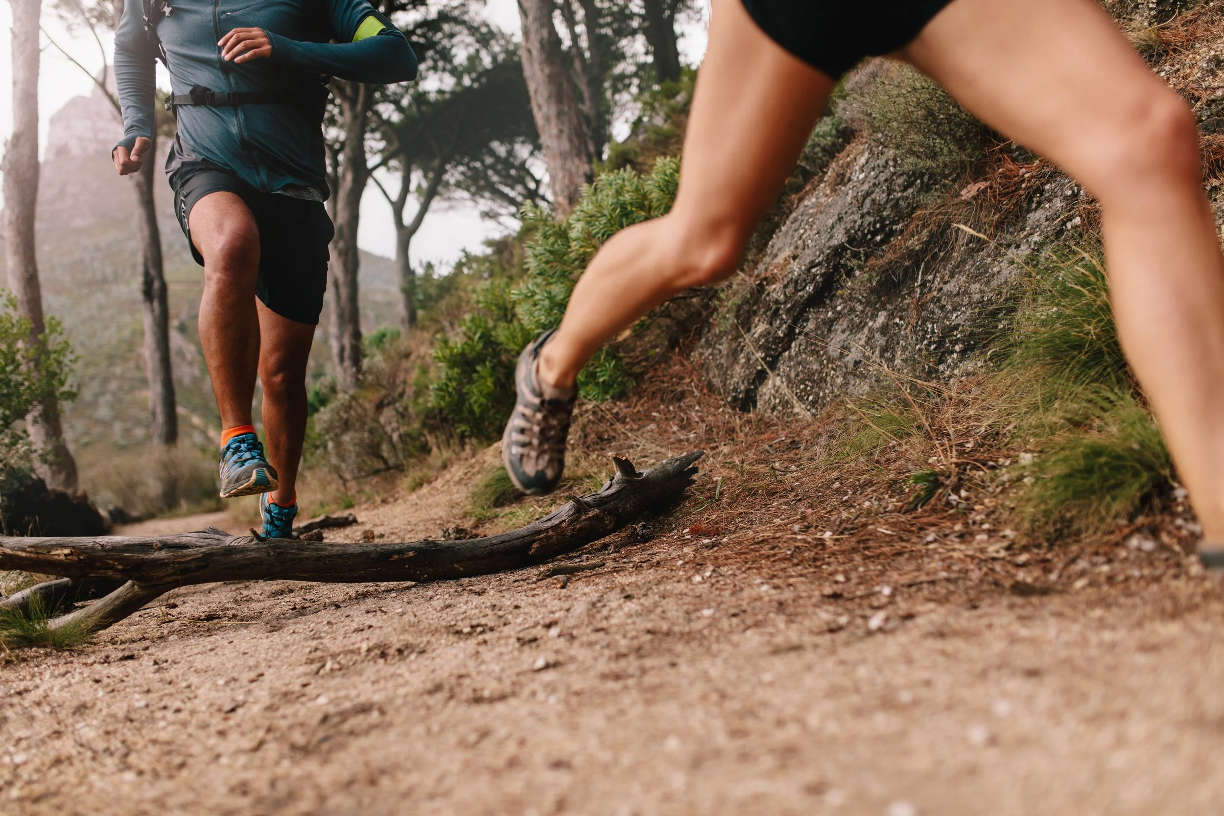 Two people trail running on a dirt trail through a forest, with a focus on their legs and shoes, one wearing black shoes and the other wearing blue shoes, amid trees and natural terrain.