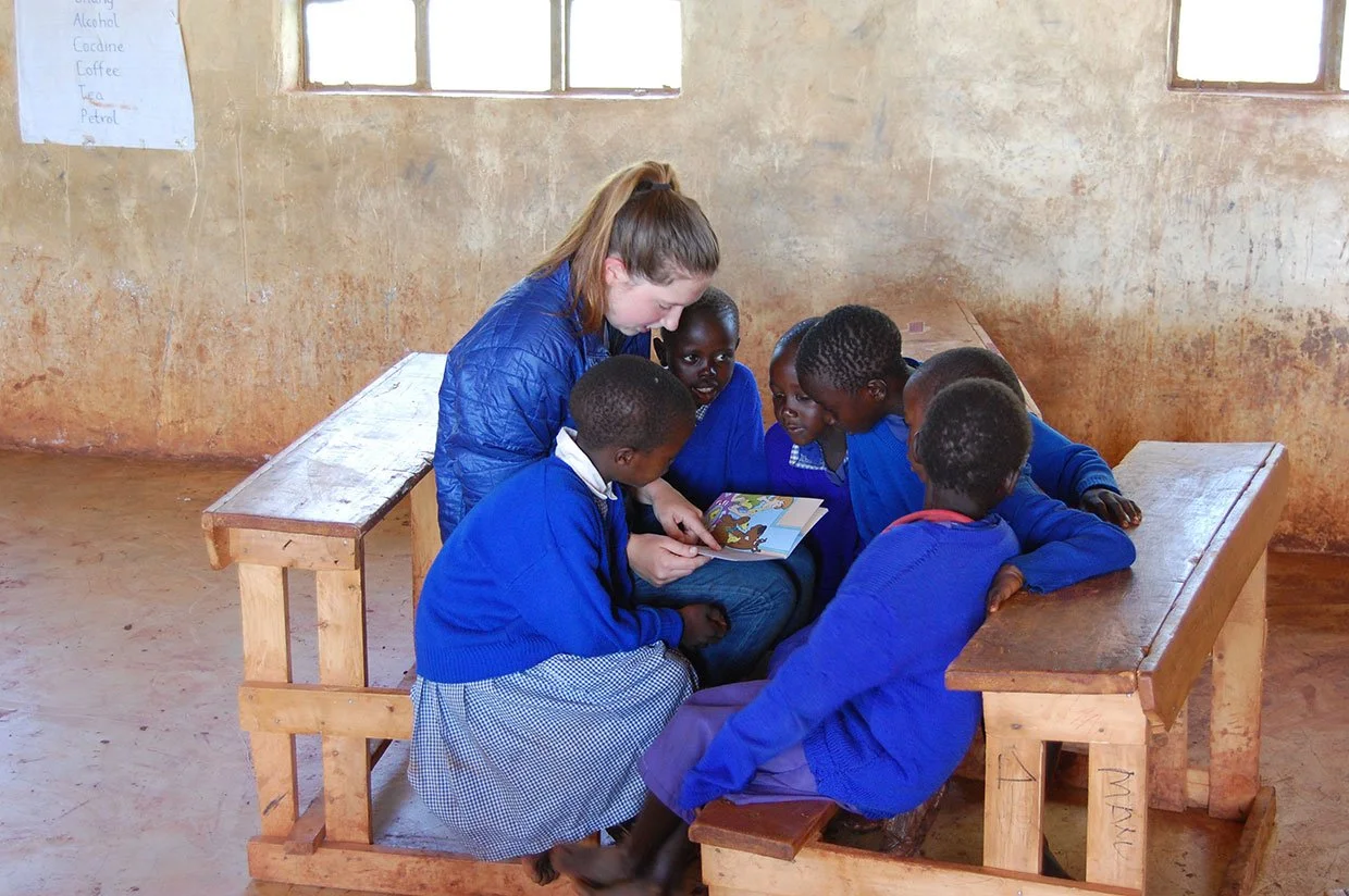 A woman and six children sitting on benches in a classroom, with the woman reading a book to the children.