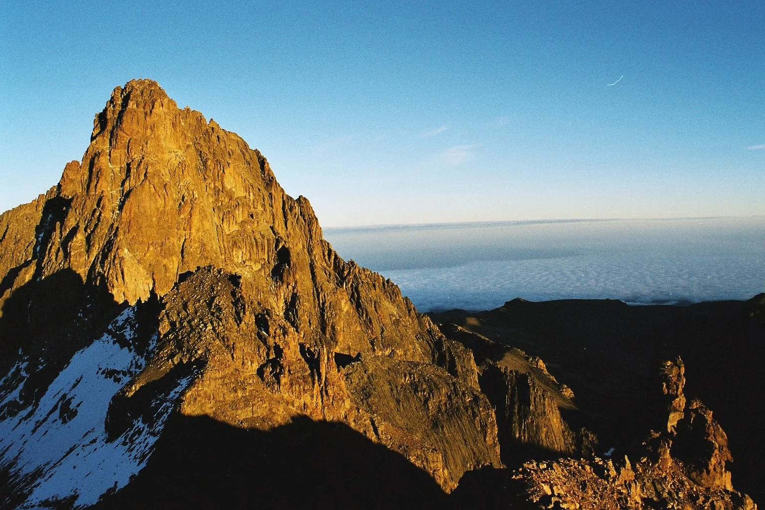A rocky mountain peak illuminated by sunlight with snow patches, overlooking a sea of clouds on the horizon under a clear blue sky.