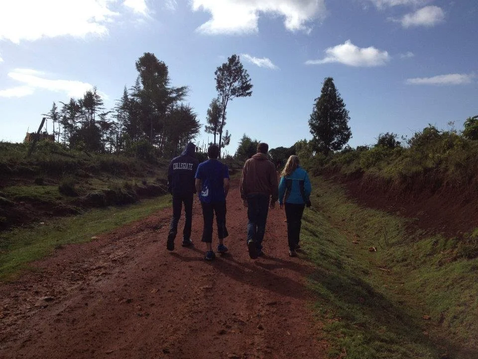 Four people walk along a dirt path in a rural area with grass and trees, under a partly cloudy sky.