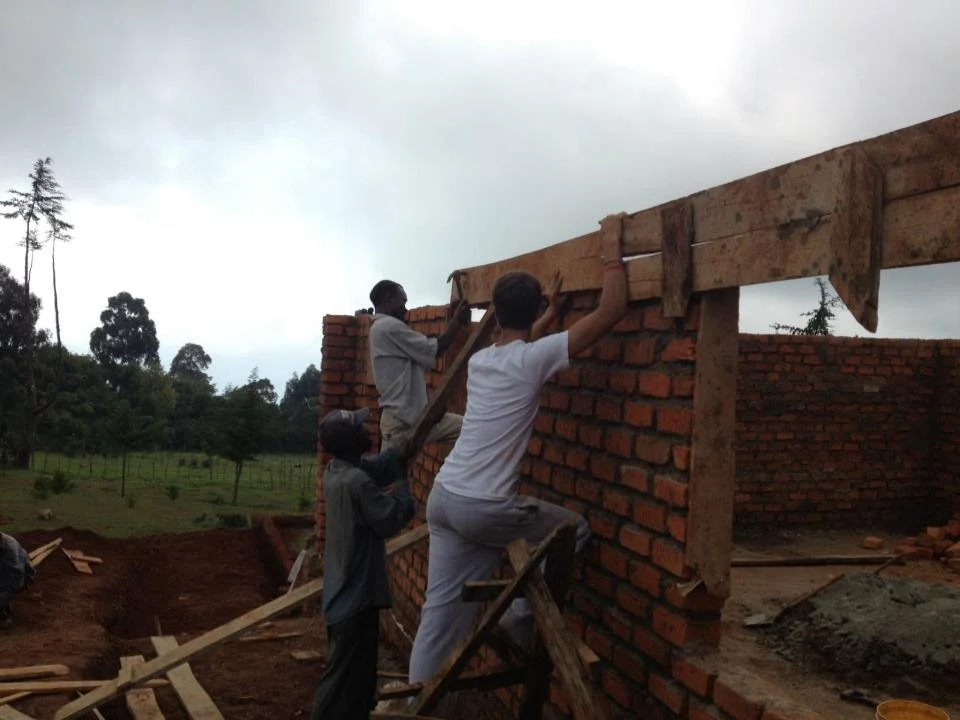 Three men building a brick wall outdoors on a cloudy day, with trees and open land in the background.
