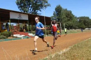 Two runners competing on a dirt track in a park, with trees and a small shelter in the background.