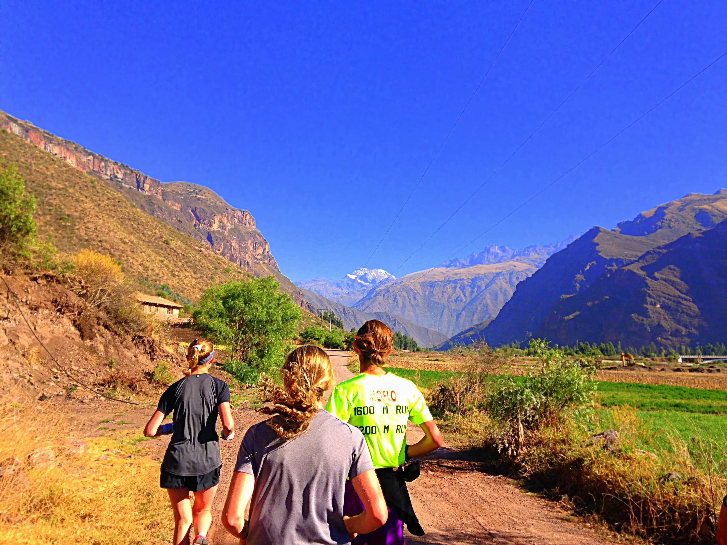 Three people jogging on a dirt trail through a lush green valley surrounded by mountains under a clear blue sky.