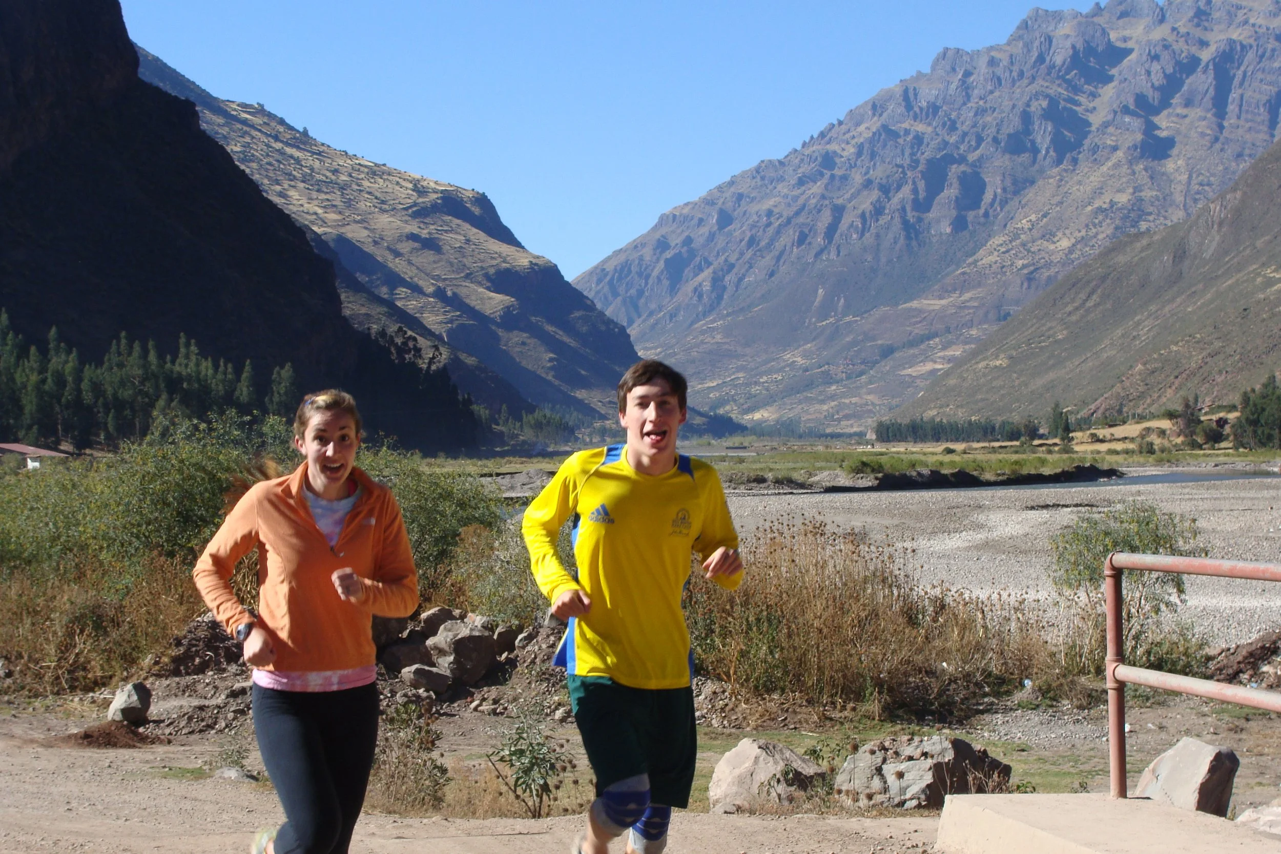 Two runners, a woman in an orange jacket and a man in a yellow shirt, running outdoors in a mountainous landscape with clear blue sky, rugged mountains, and sparse vegetation.