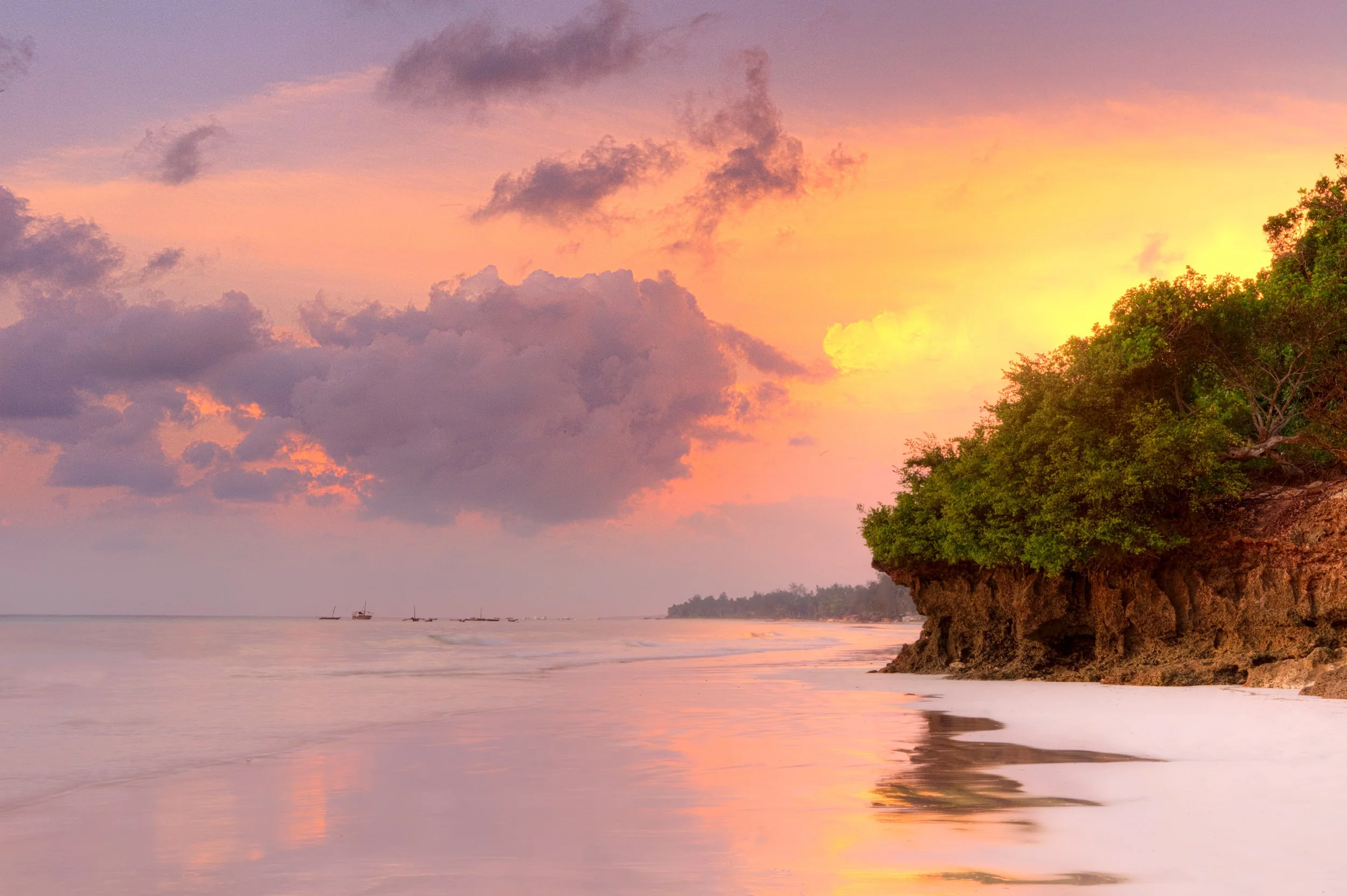 Sunset over a beach with a rocky outcrop covered in green trees on the right, partly cloudy sky with pink, purple, and orange hues, gentle waves, and boats in the distance.