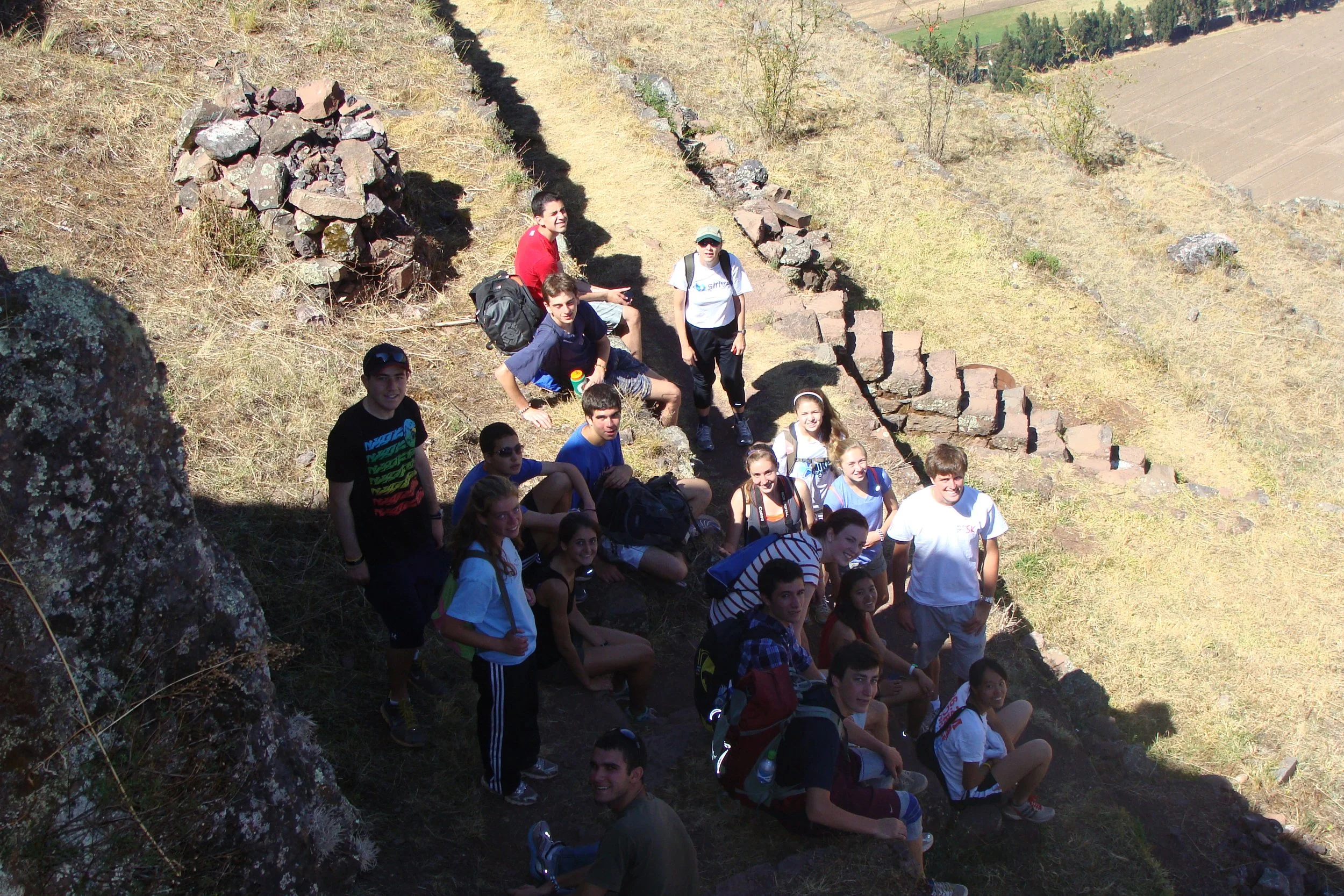 Group of young people on a hiking trip resting on a hillside with stone ruins and a scenic valley in the background.