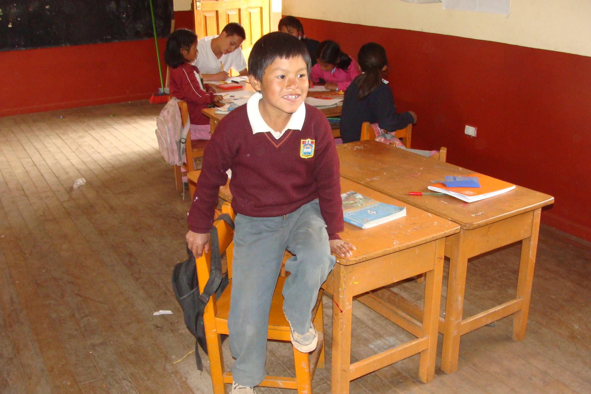 A young boy in a school uniform climbing on a chair in a classroom with other students working at desks in the background.