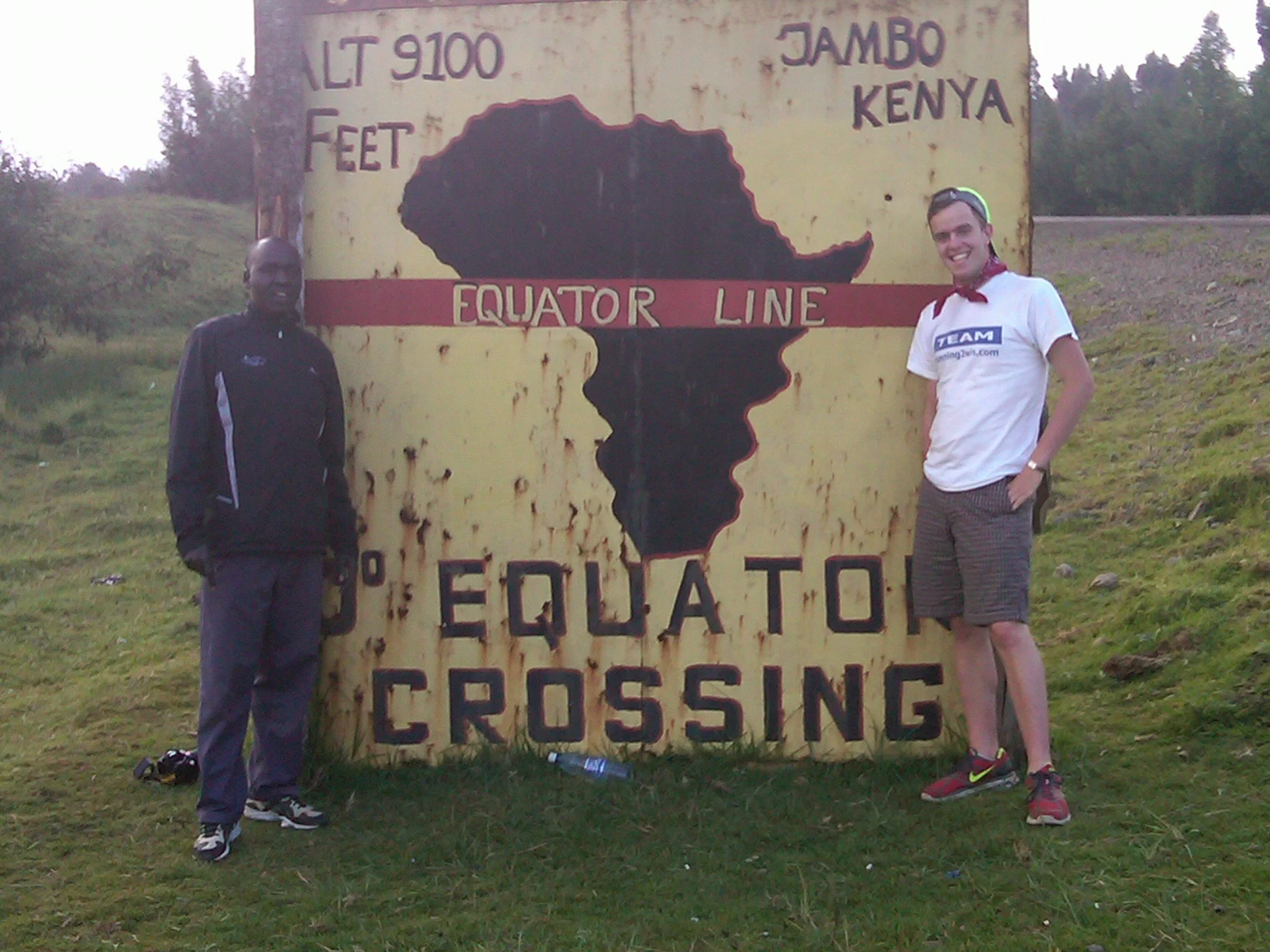 Two people standing near a large sign that reads 'Equator Crossing,' with a map of Africa depicting Kenya and the words 'Jambo Kenya' and 'Lt 9100 feet.' One person is wearing a black jacket and gray pants, and the other is wearing a white t-shirt, s