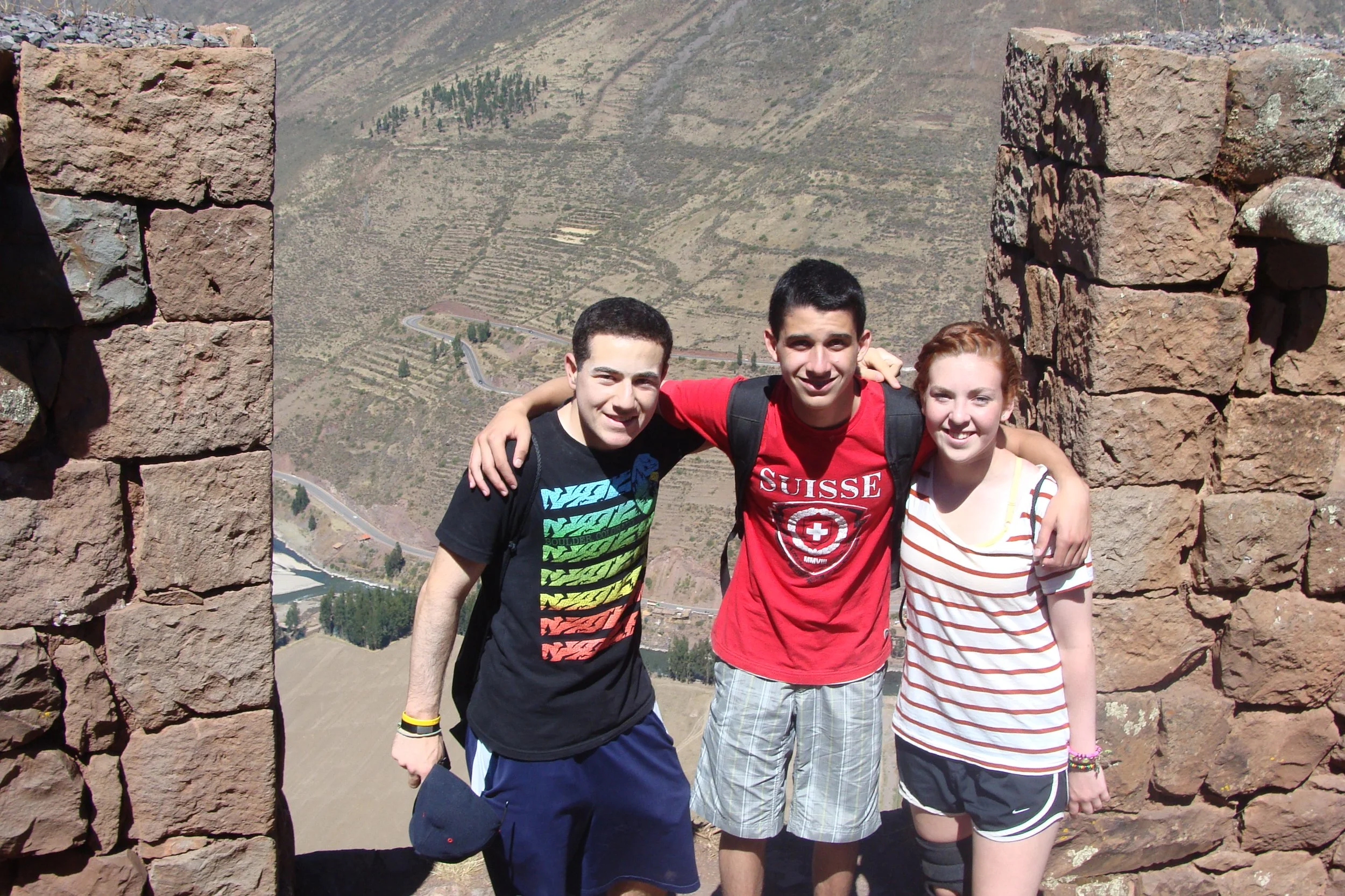 Three young friends standing between two stone brick walls on a mountain overlooking a winding road and terraced hills in the background.