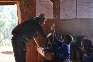 An older man in a black shirt and white cap giving a high-five to a young girl in a blue jacket inside a classroom. Other children are visible sitting at desks, and educational posters are on the wall.