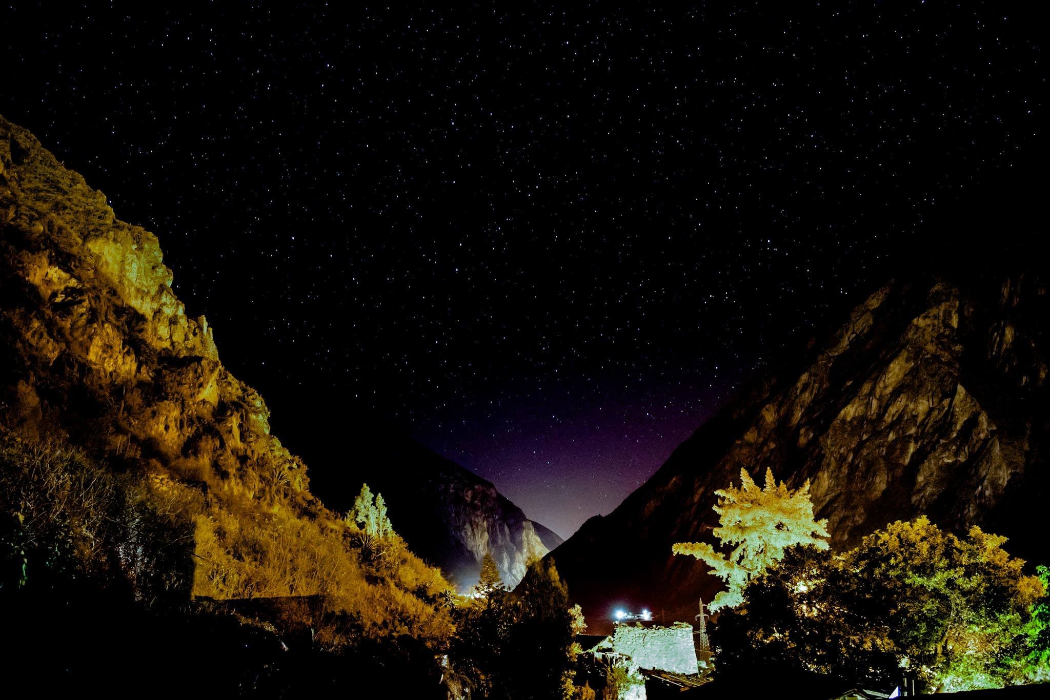 Nighttime view of a starry sky over a valley surrounded by mountains, with illuminated trees and a building in the foreground.