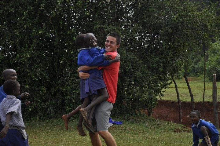A man is holding and lifting a smiling young girl in a blue school uniform. They are outdoors with trees and grass in the background. Several other children are nearby.