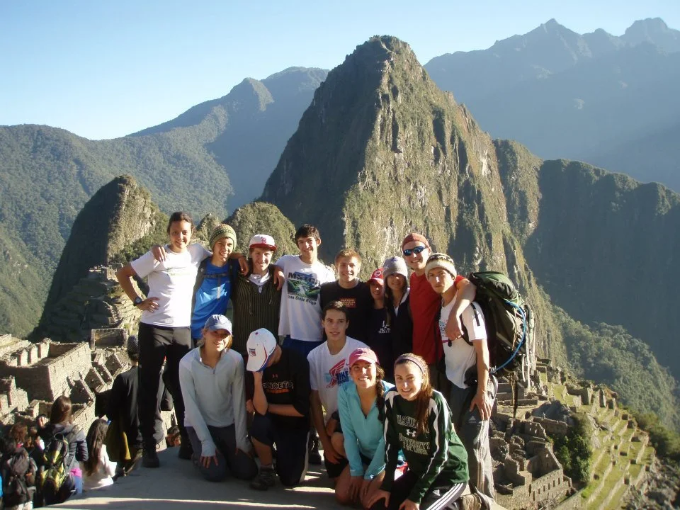 A group of about fifteen young tourists posing in front of Machu Picchu mountain in Peru, with the ancient ruins and terraces visible in the background.