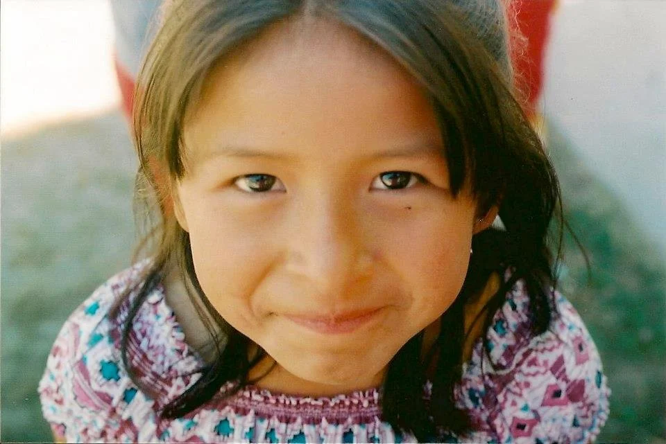 Close-up of a young girl with dark hair, smiling softly, wearing a colorful embroidered shirt, outdoors with blurred background.