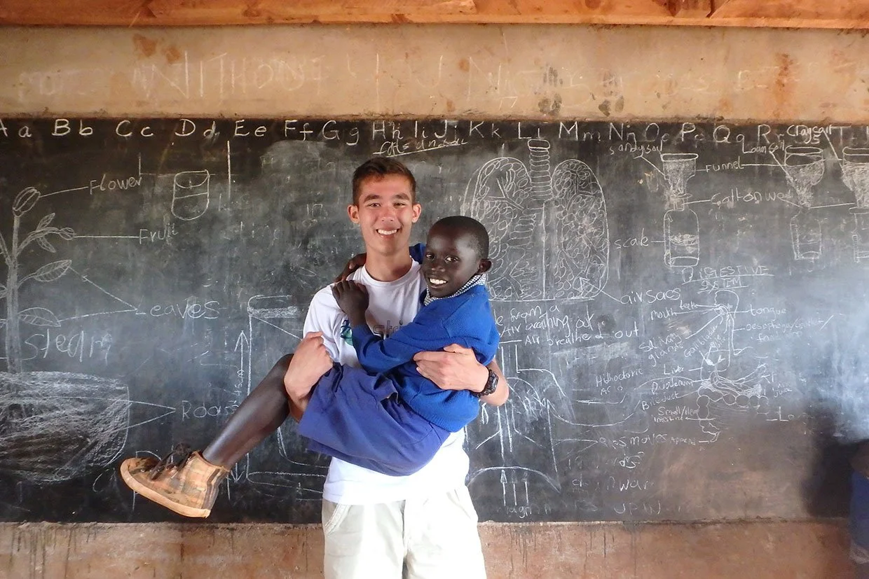 A young man in a white shirt holding a smiling little boy in a blue uniform in front of a chalkboard with diagrams and labels of the human digestive system and plant parts.