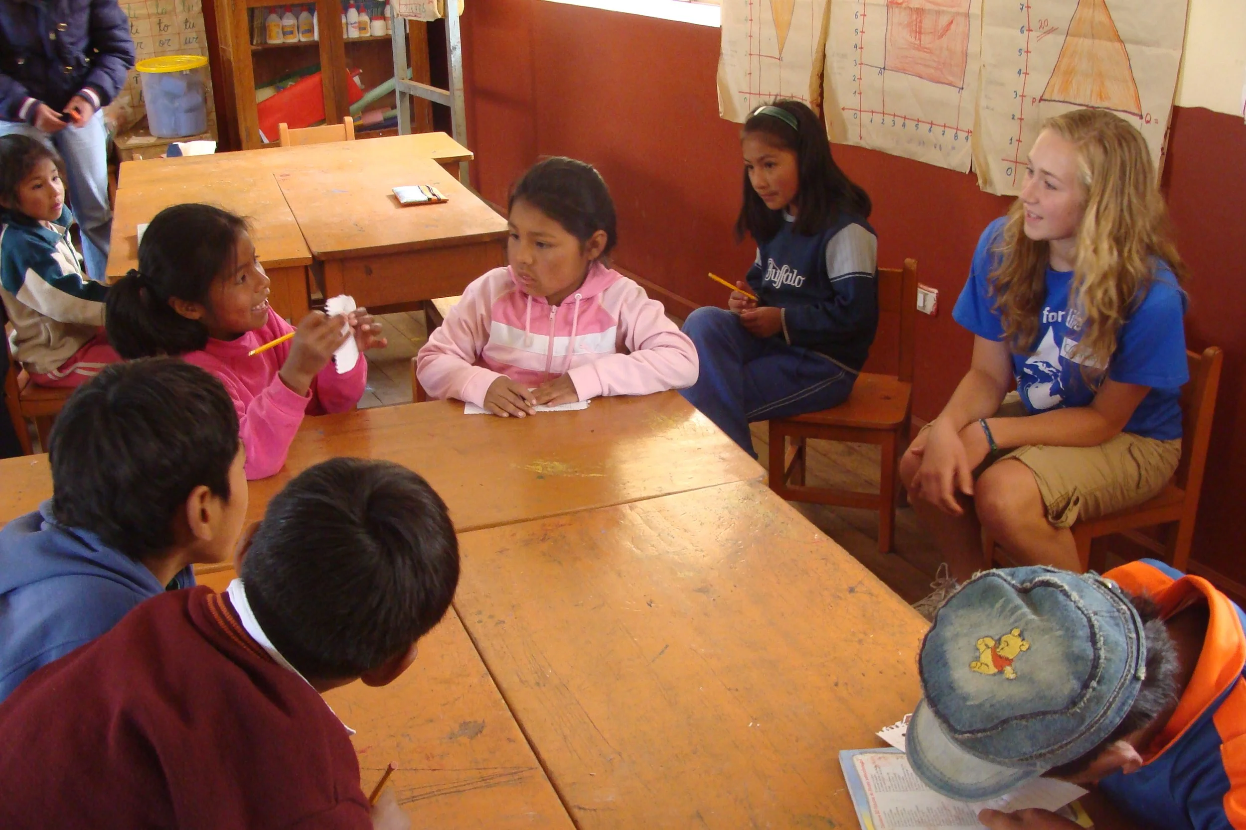 Children and a young woman sitting at a wooden table in a classroom, engaging in a discussion or activity. Some children are holding pencils, and there are educational posters on the wall.