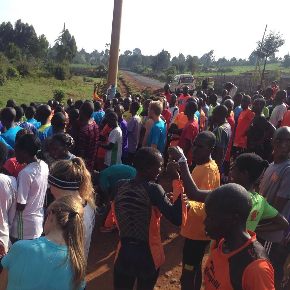 A group of people, mostly African, gathered outdoors on a dirt road with greenery and trees in the background. Some are talking, while others are looking towards a person holding a flag or banner. A vehicle is parked nearby.