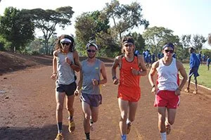 Four people running outdoors on a dirt path, dressed in athletic wear, with trees and a clear sky in the background.