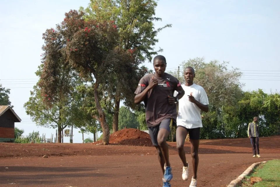 Two young men running outdoors on a dirt path with trees and a house in the background.