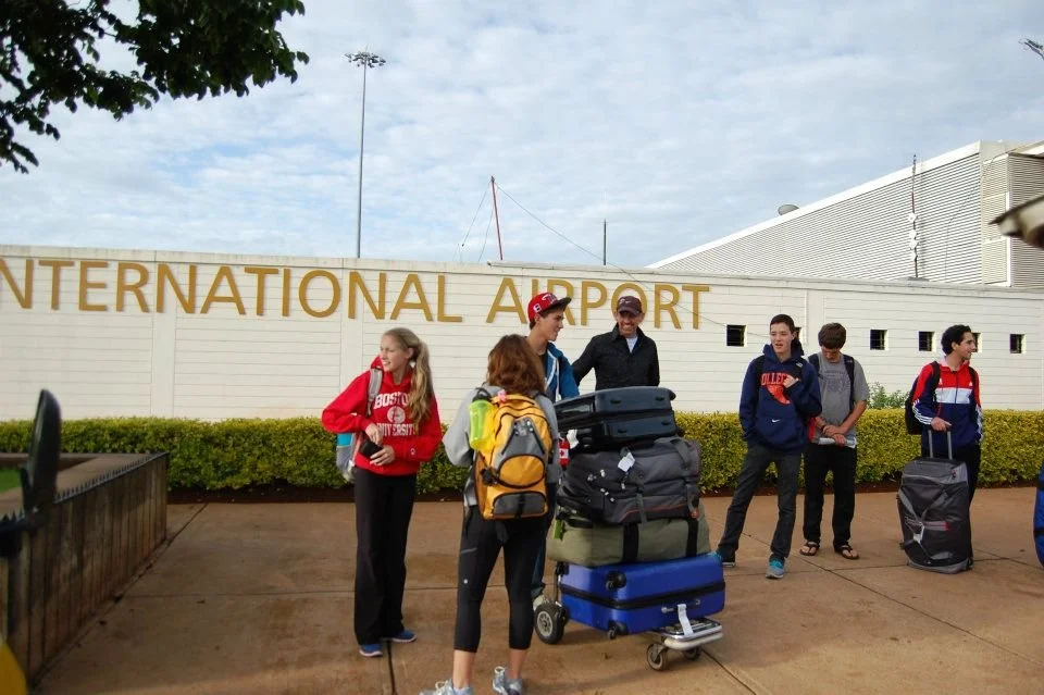 Group of young travelers with luggage standing outside the entrance of an airport, with 'INTERNATIONAL AIRPORT' sign in the background.
