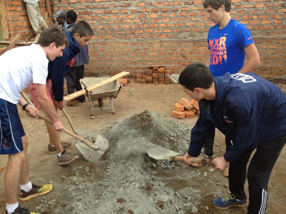 Group of young men working together to move a pile of dirt and rocks with shovels at a construction site, with bricks and a wheelbarrow nearby.
