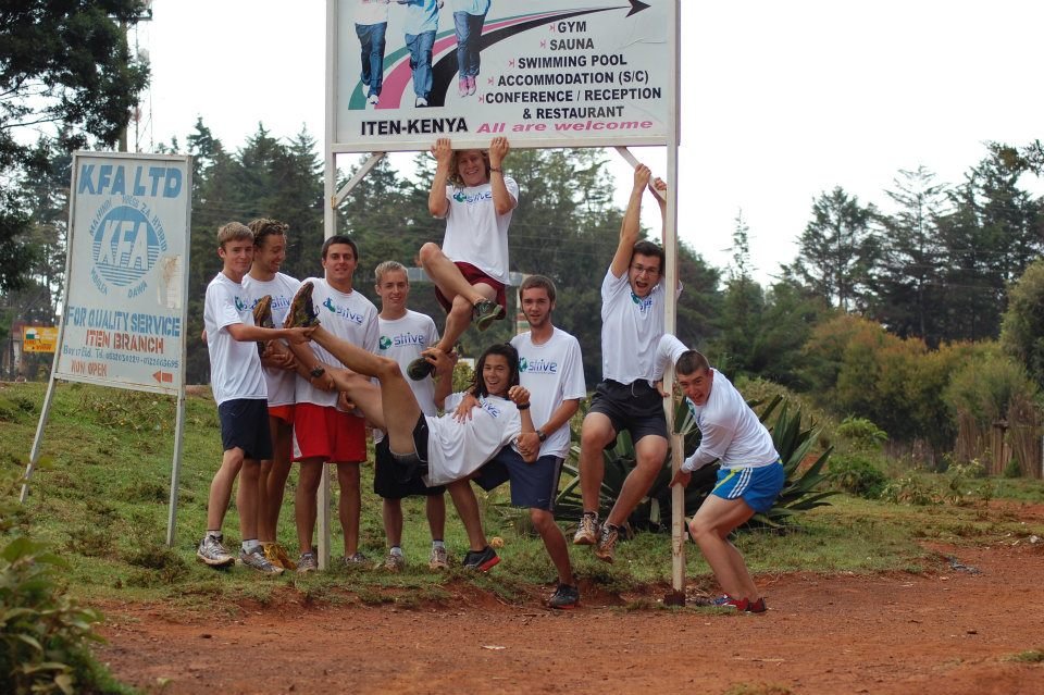 A group of seven young people in white t-shirts and shorts pose outdoors, holding a small wooden sign and supporting a person sitting on their shoulders near a large signboard with directions to various facilities.