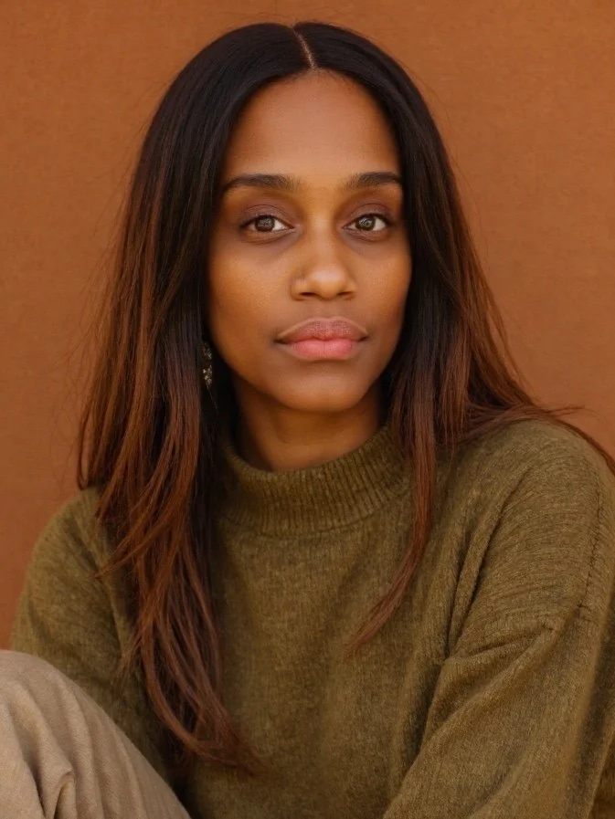 A young woman with long brown hair and a neutral expression, wearing an olive green sweater, in front of a plain brown background.