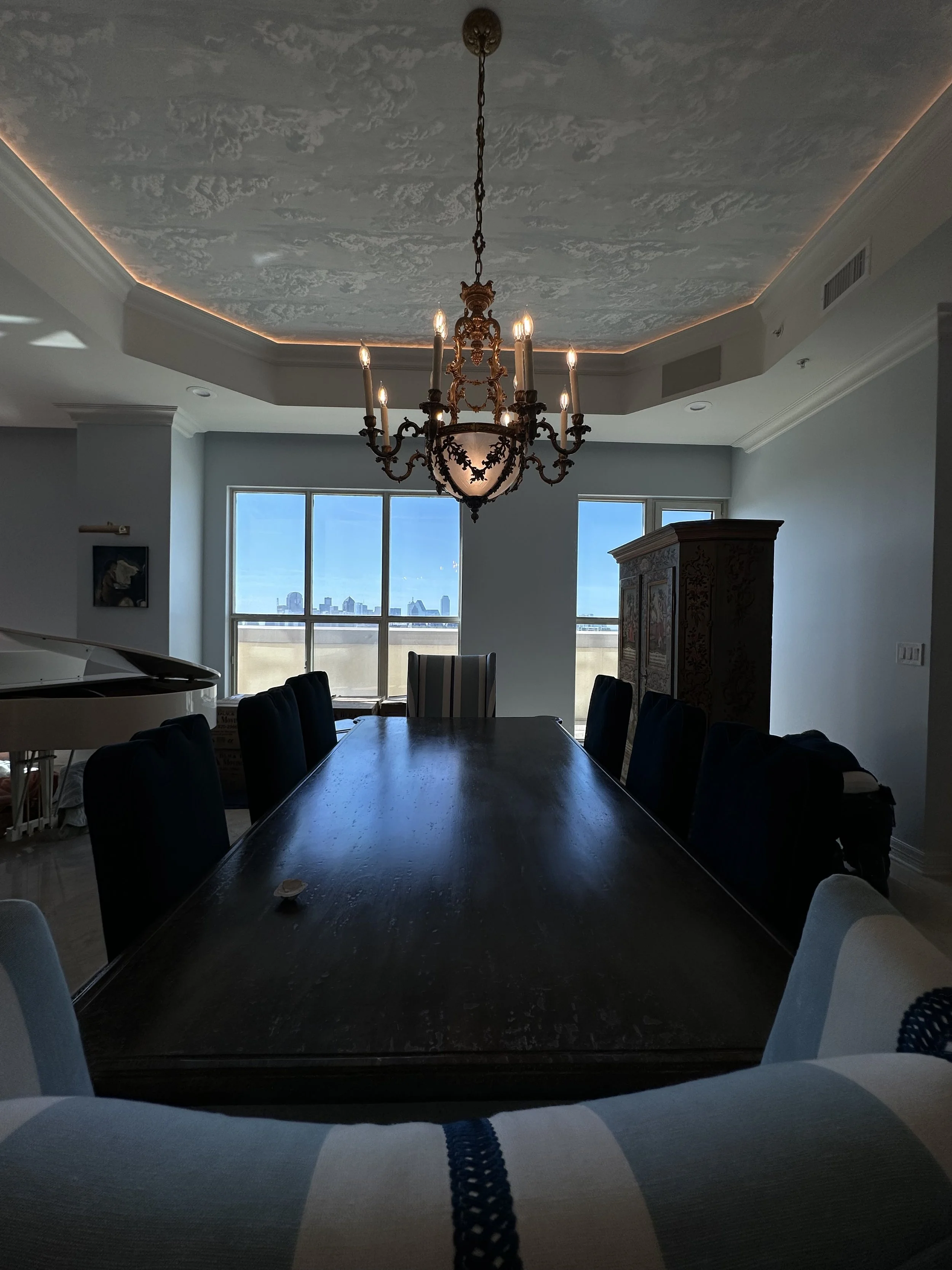A dining room with a large wooden table, black and white chairs, a chandelier hanging from the ceiling, and a large window showing a city skyline in the background.