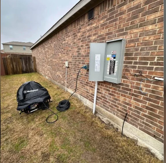 Outdoor scene with an electrical panel mounted on a brick wall, connected to a black cable, on a grassy yard with a black object nearby and a wooden fence in the background.