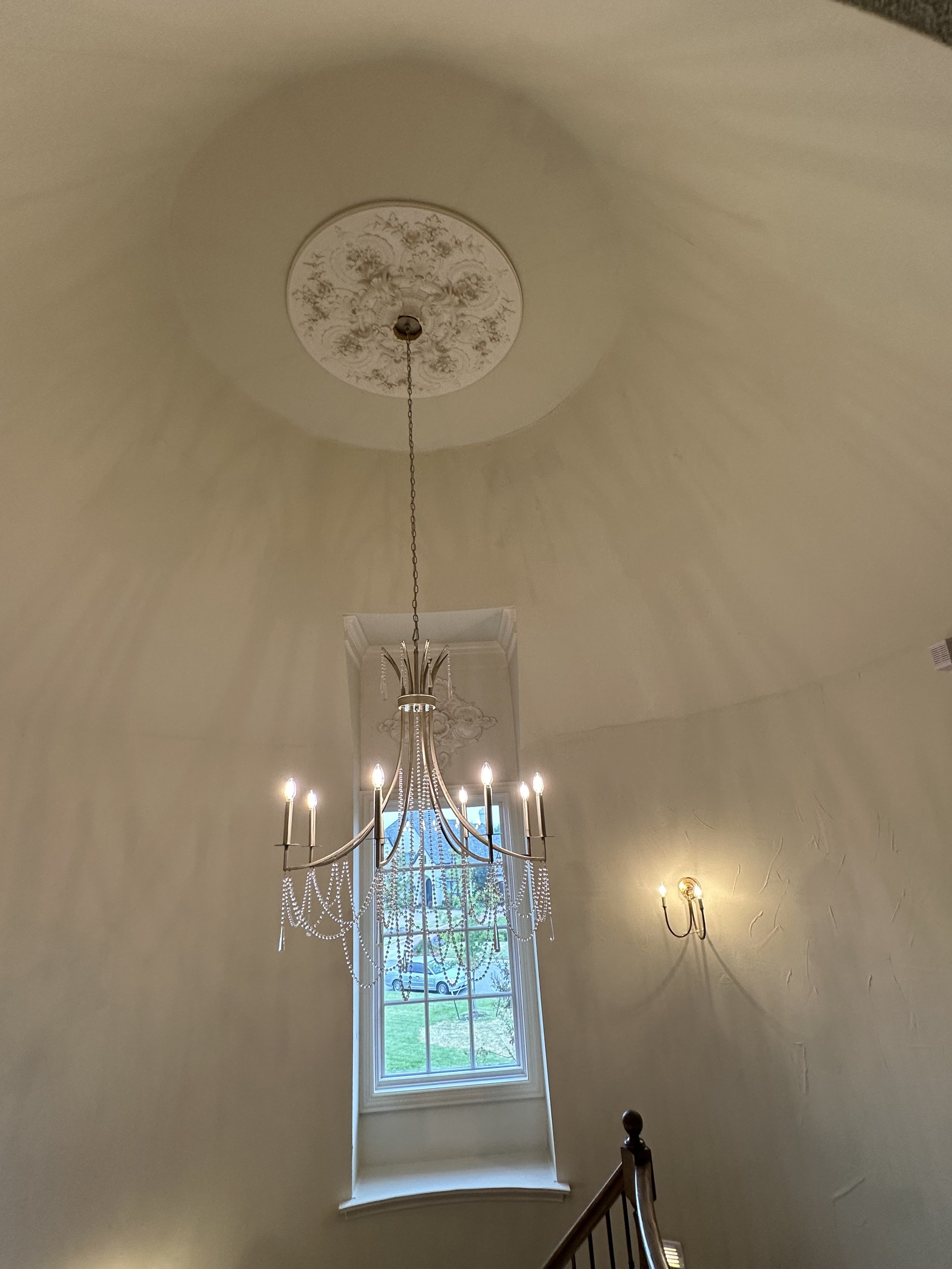 Chandelier hanging from a decorative ceiling medallion in a tall, curved-ceiling foyer with a window showing a lawn and parked car outside.