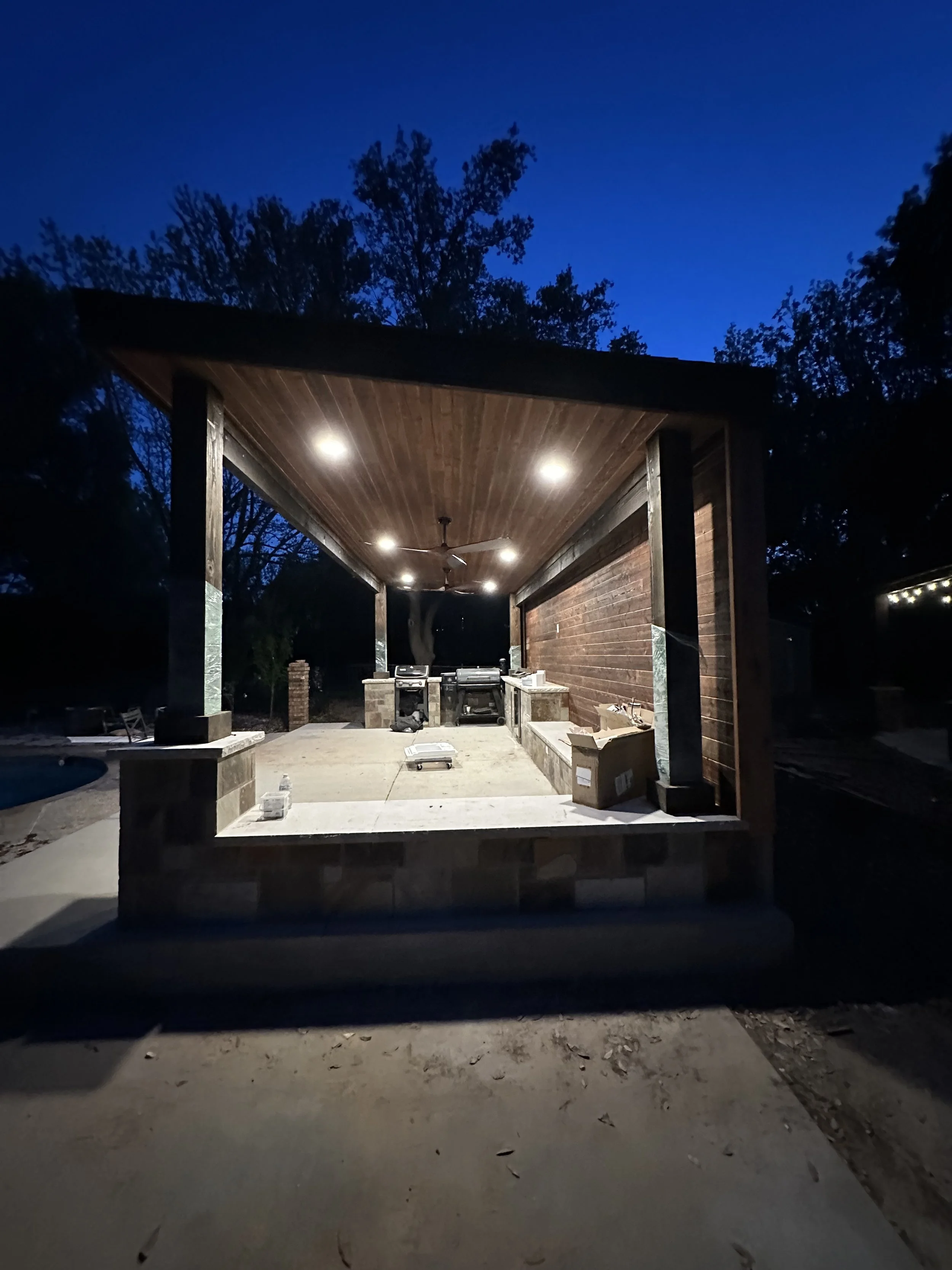 Outdoor kitchen area with a wooden ceiling and brick walls, illuminated by ceiling lights, featuring a built-in grill, countertop, and some boxes and tools under construction, surrounded by trees at dusk.