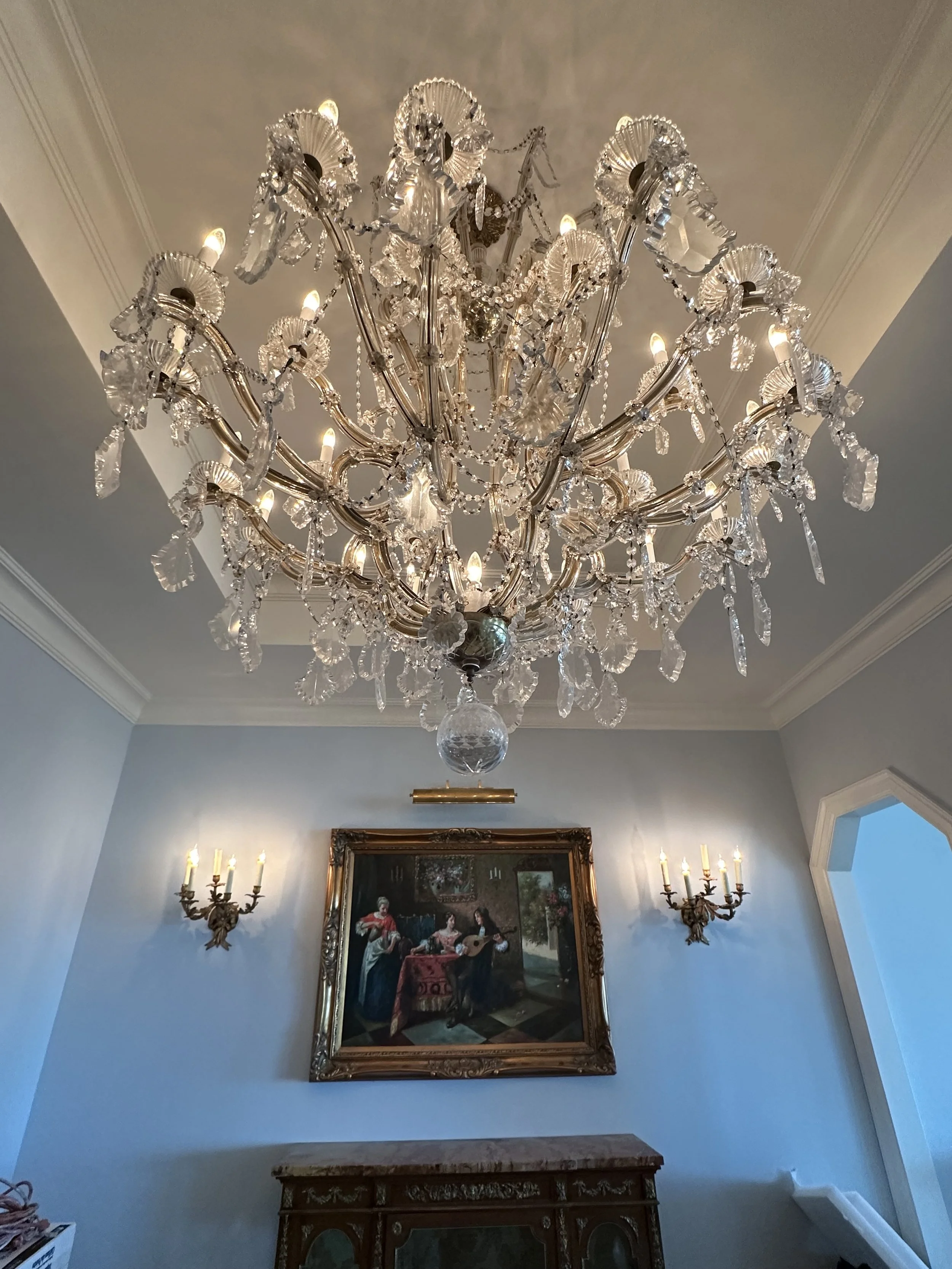 A large crystal chandelier hanging from a ceiling in a room with light blue walls, a framed painting depicting people and a guitar, and wall-mounted light fixtures.