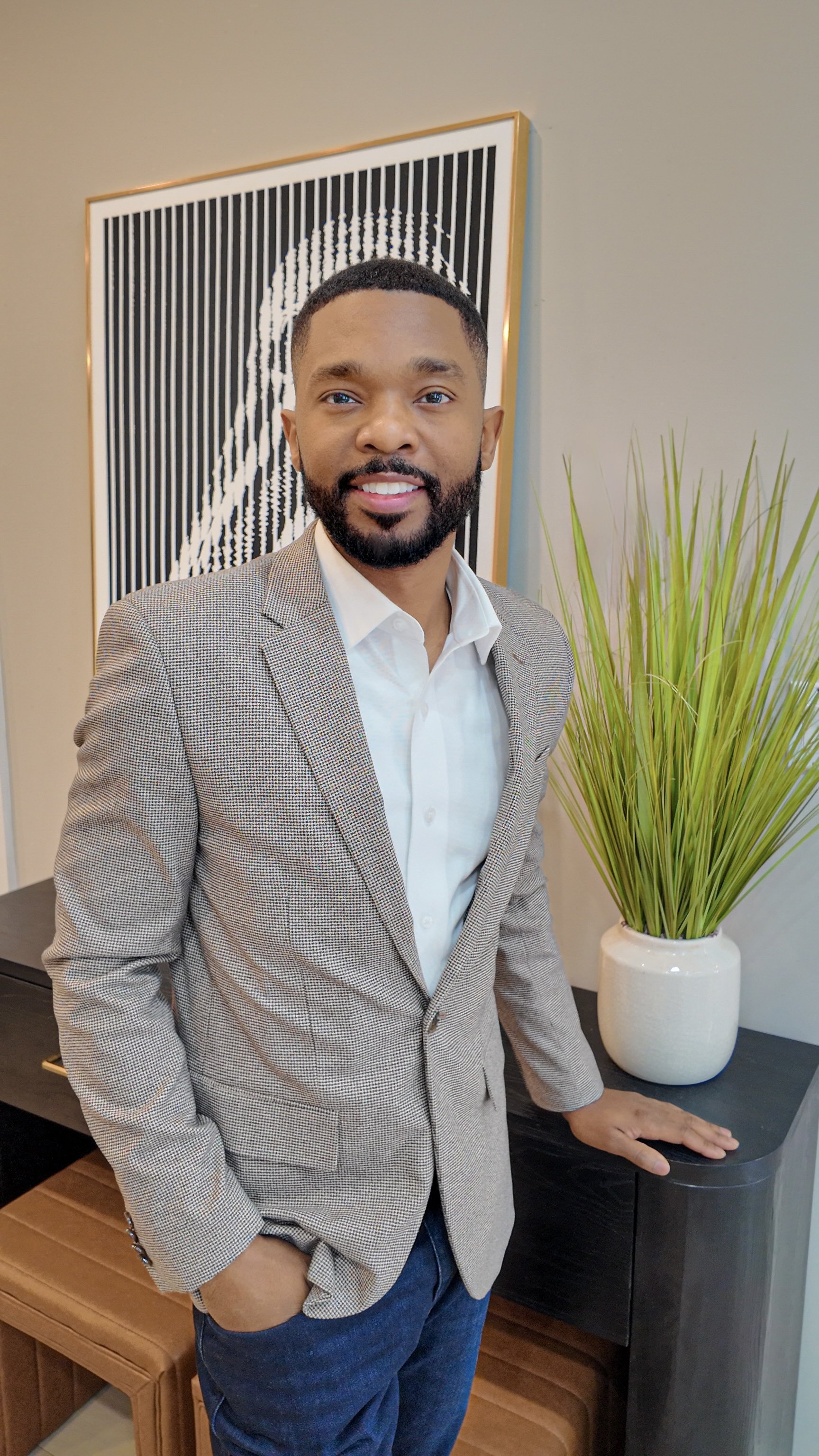A man with a beard and mustache wearing a beige checkered blazer, white shirt, and dark jeans, standing indoors beside a black table with a large white vase of green plants and a framed black and white artwork in the background.